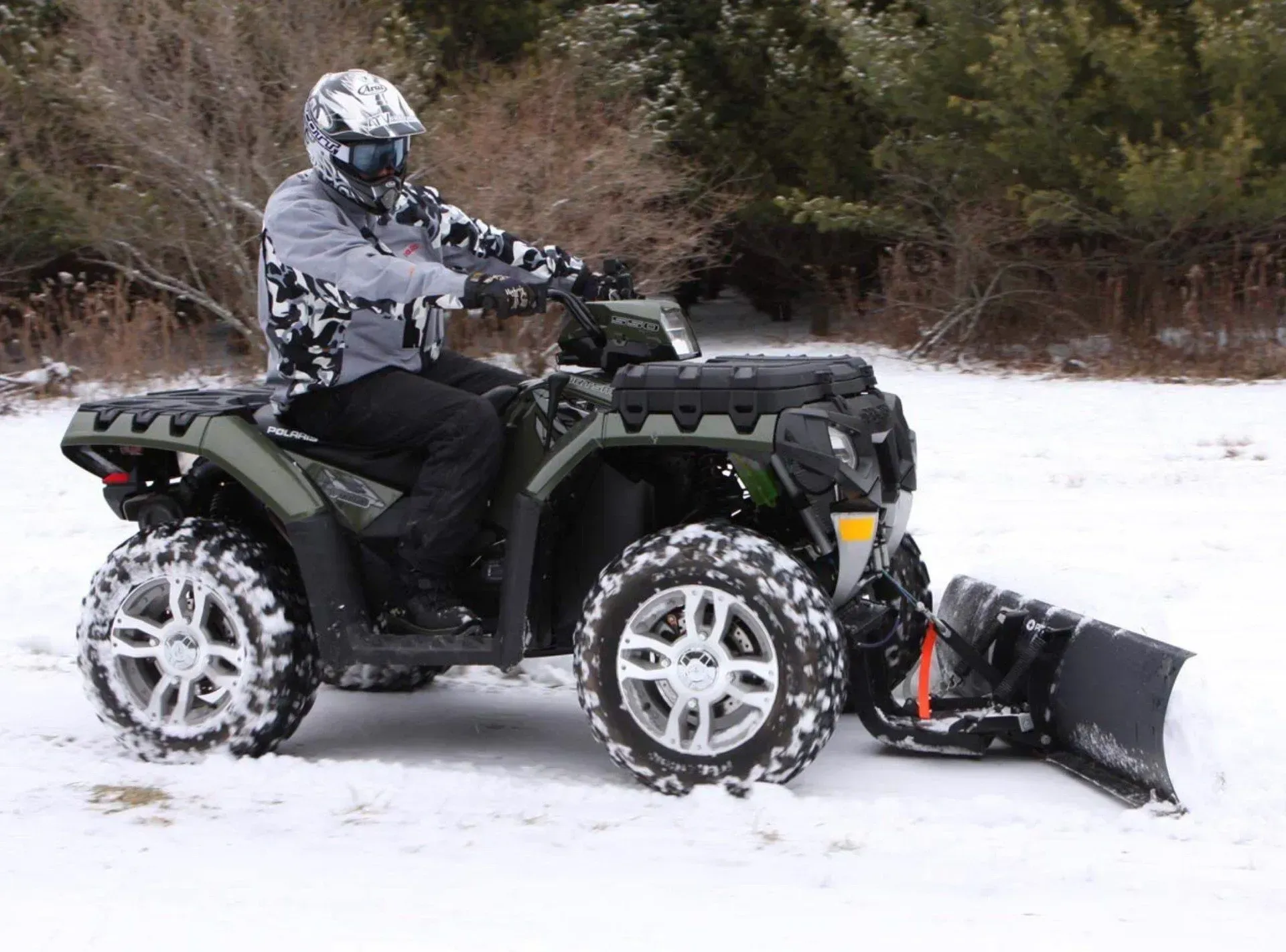 Person on a green ATV plowing snow in a snowy area; rider wearing helmet and winter gear.