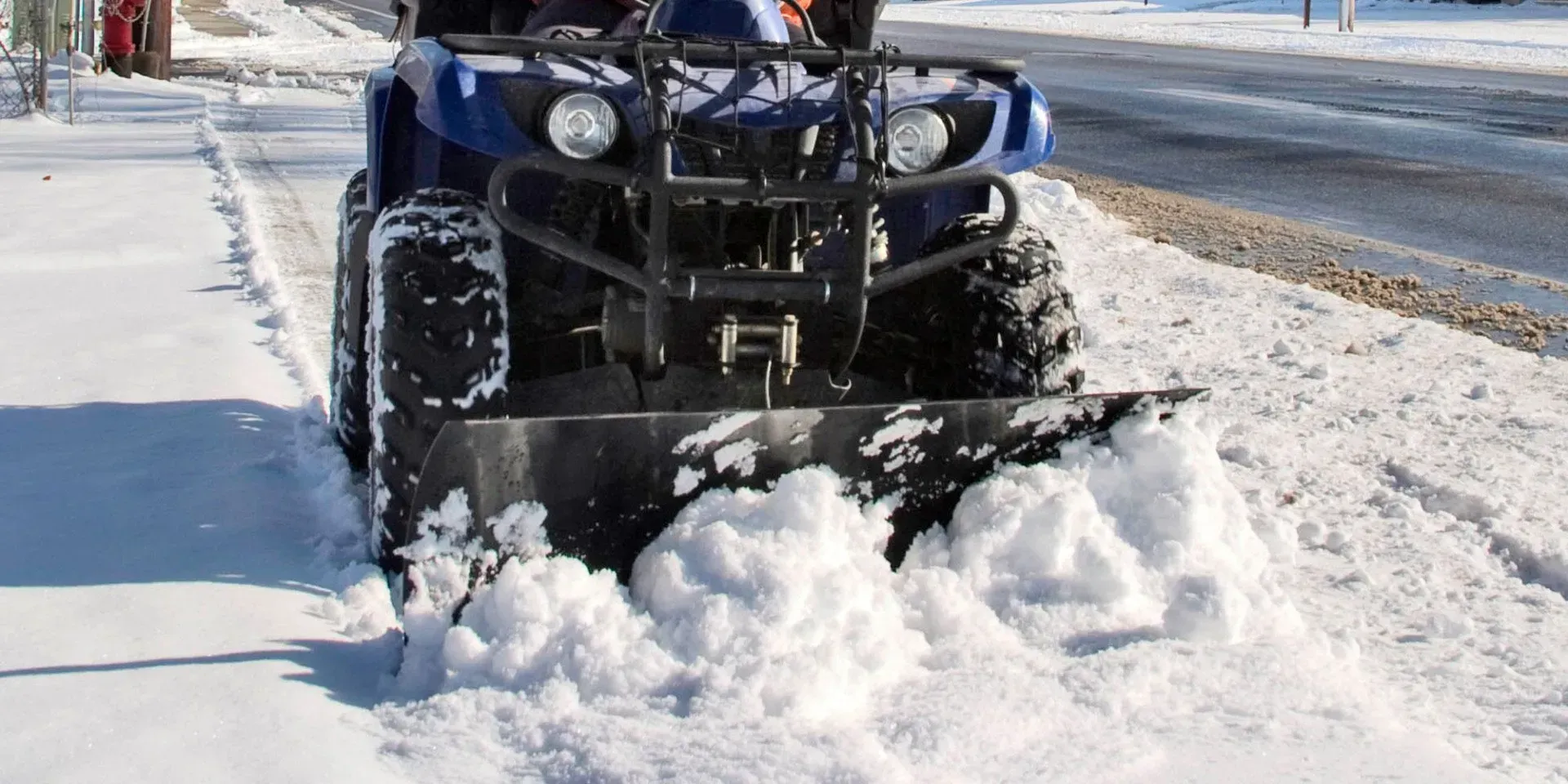 Blue ATV with a front snow plow blade clearing a snowy sidewalk